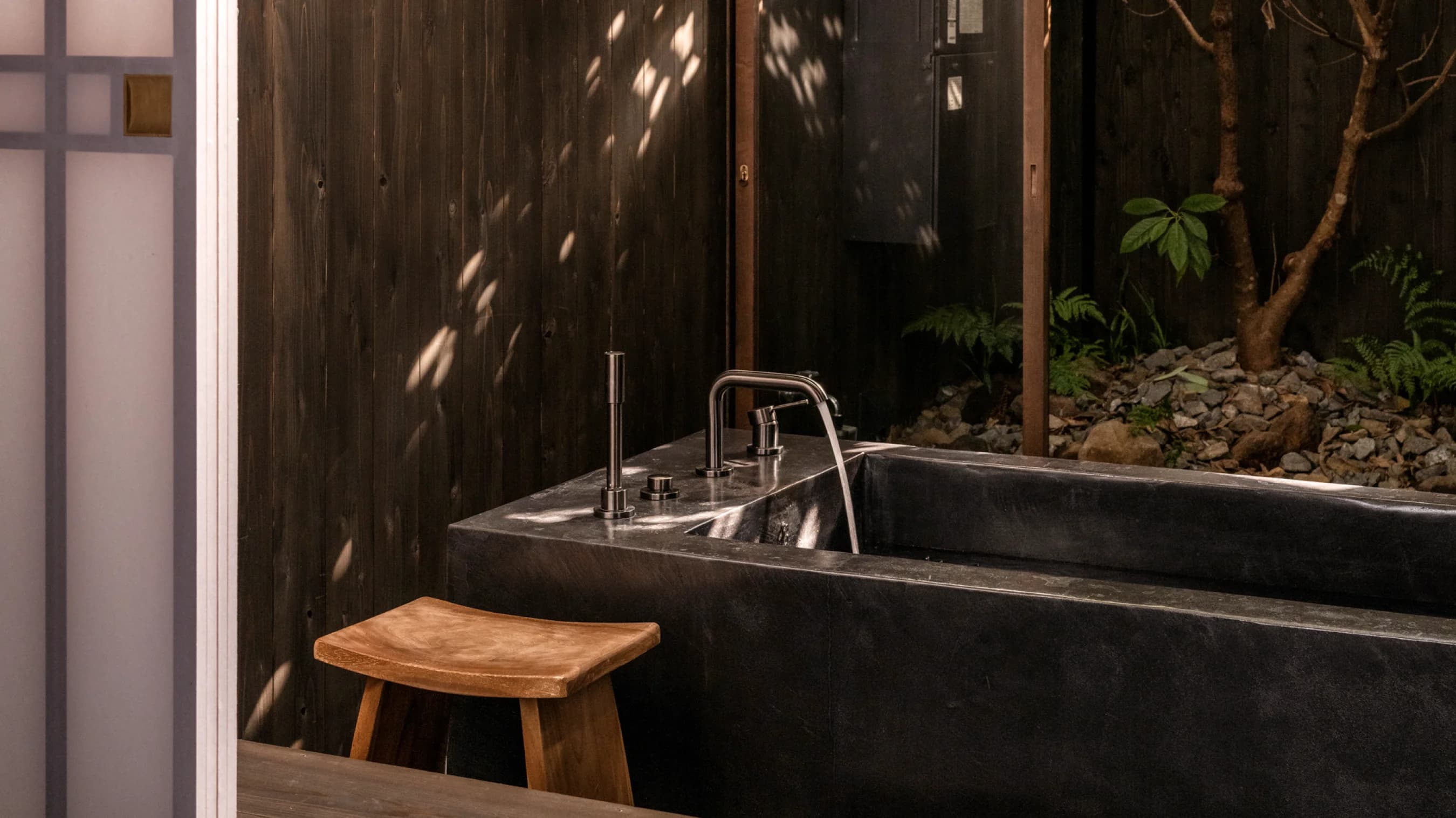 Open-air bath area with wooden stool and stone basins beside a garden
